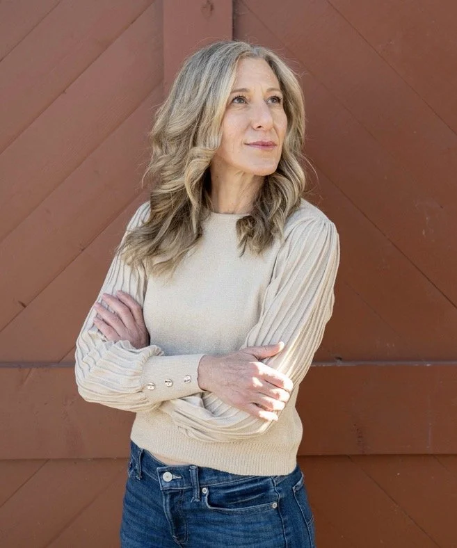A mature woman with long, wavy, blonde hair stands outdoors in front of a reddish-brown wooden wall. She is wearing a beige long-sleeve sweater and blue jeans. She crosses her arms and gazes thoughtfully into the distance.