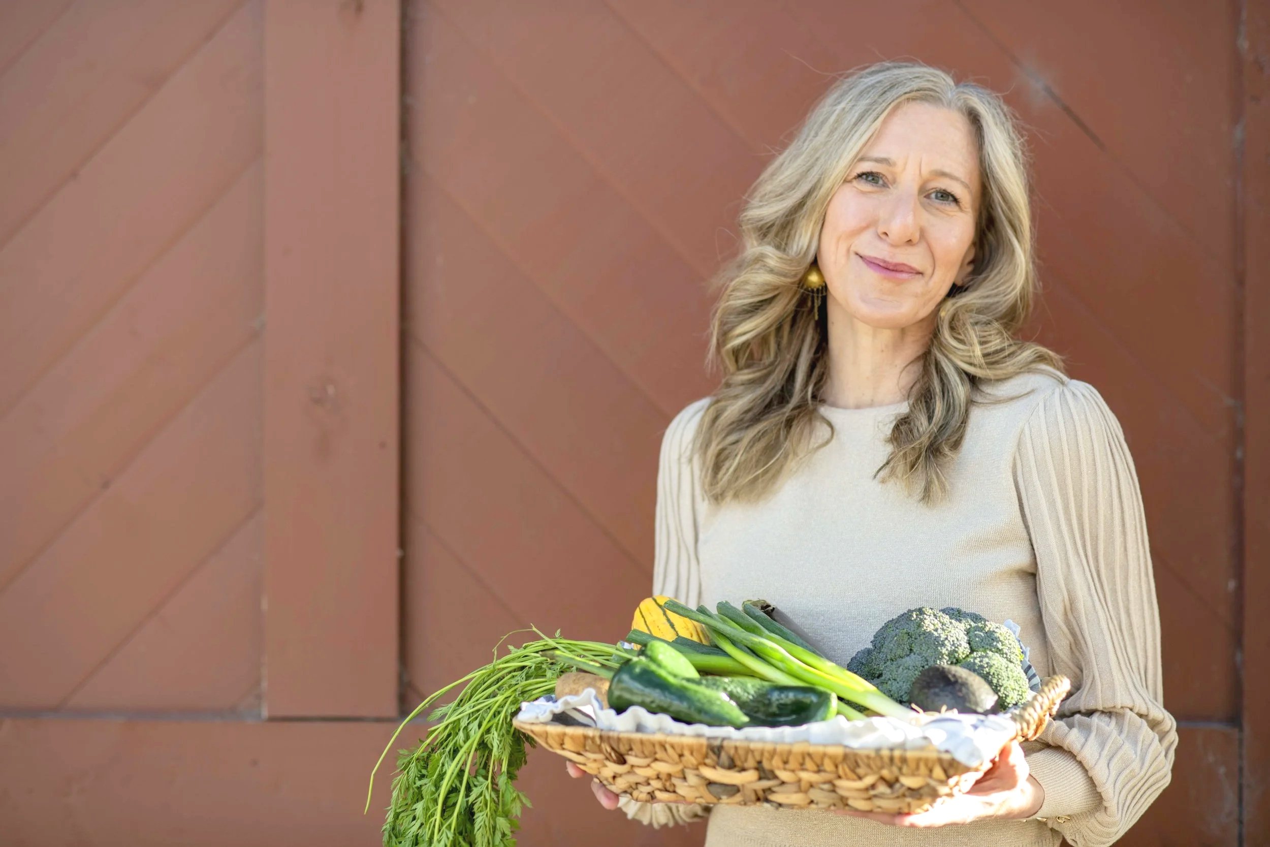 A woman with blonde hair holding a basket of fresh vegetables, including zucchini, broccoli, and carrots, standing outdoors against a brown wooden fence.