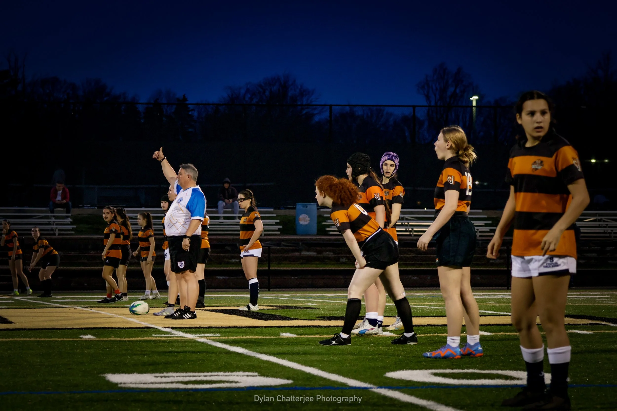 A group of female rugby players in black and orange uniforms on an outdoor field during evening, with a referee and spectators in the background.