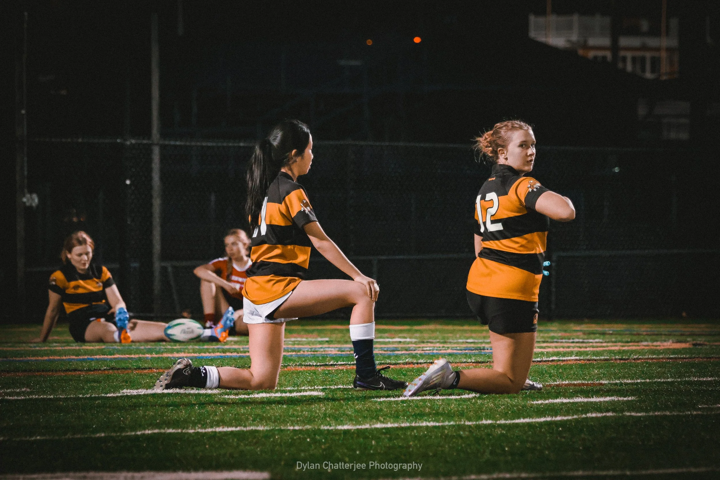 Girls in sports uniforms kneeling and stretching on a synthetic turf field at night, with a fence and dark sky in the background.