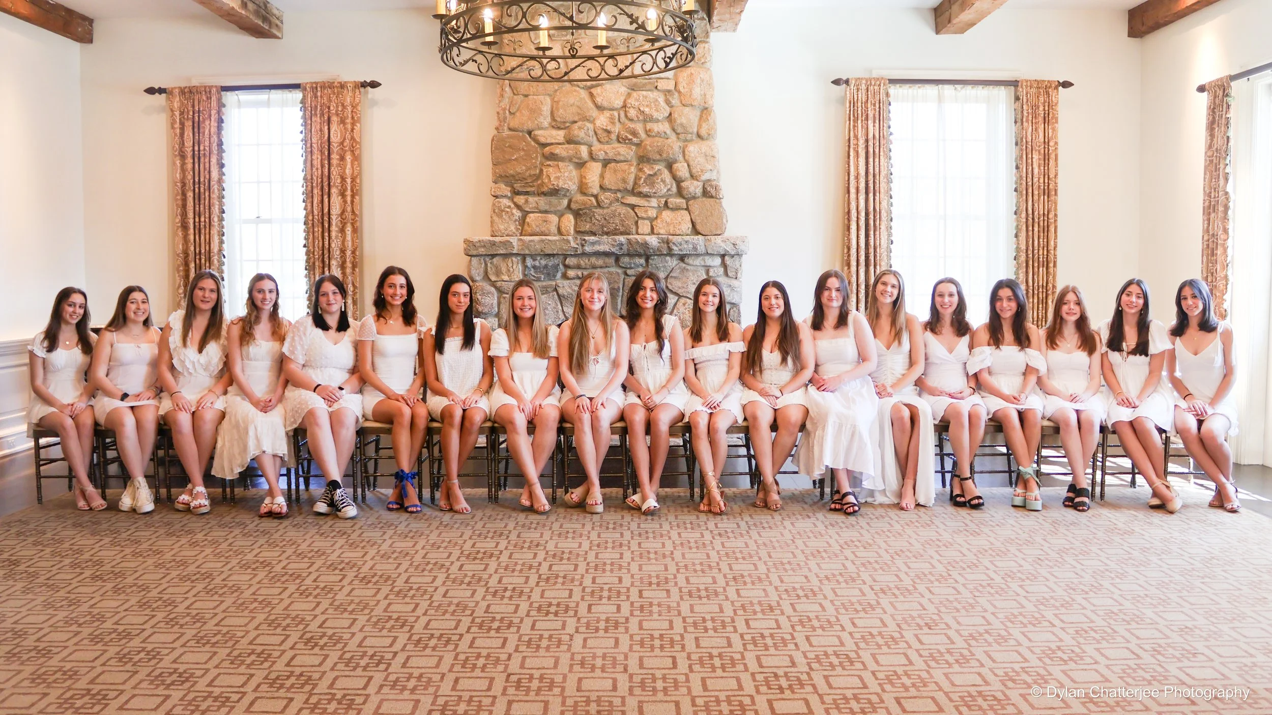 Group of young women seated in a line, dressed in white, in a bright room with large windows, stone fireplace, and chandelier.