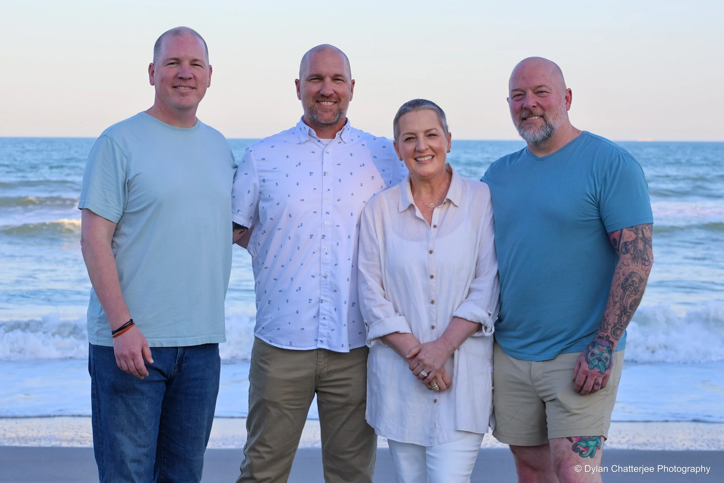 Four people standing on a beach near the water, smiling at the camera, with the ocean and sky in the background.