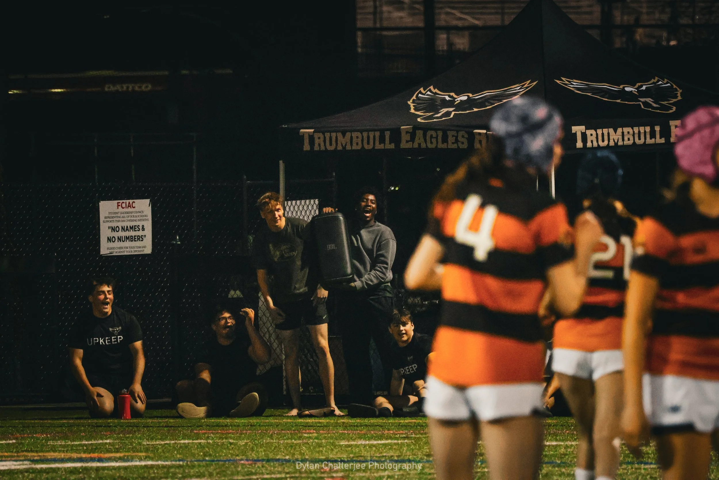 People watching a football game at night, with the focus on players. Some spectators are sitting and cheering in front of a Trumbull Eagles tent.