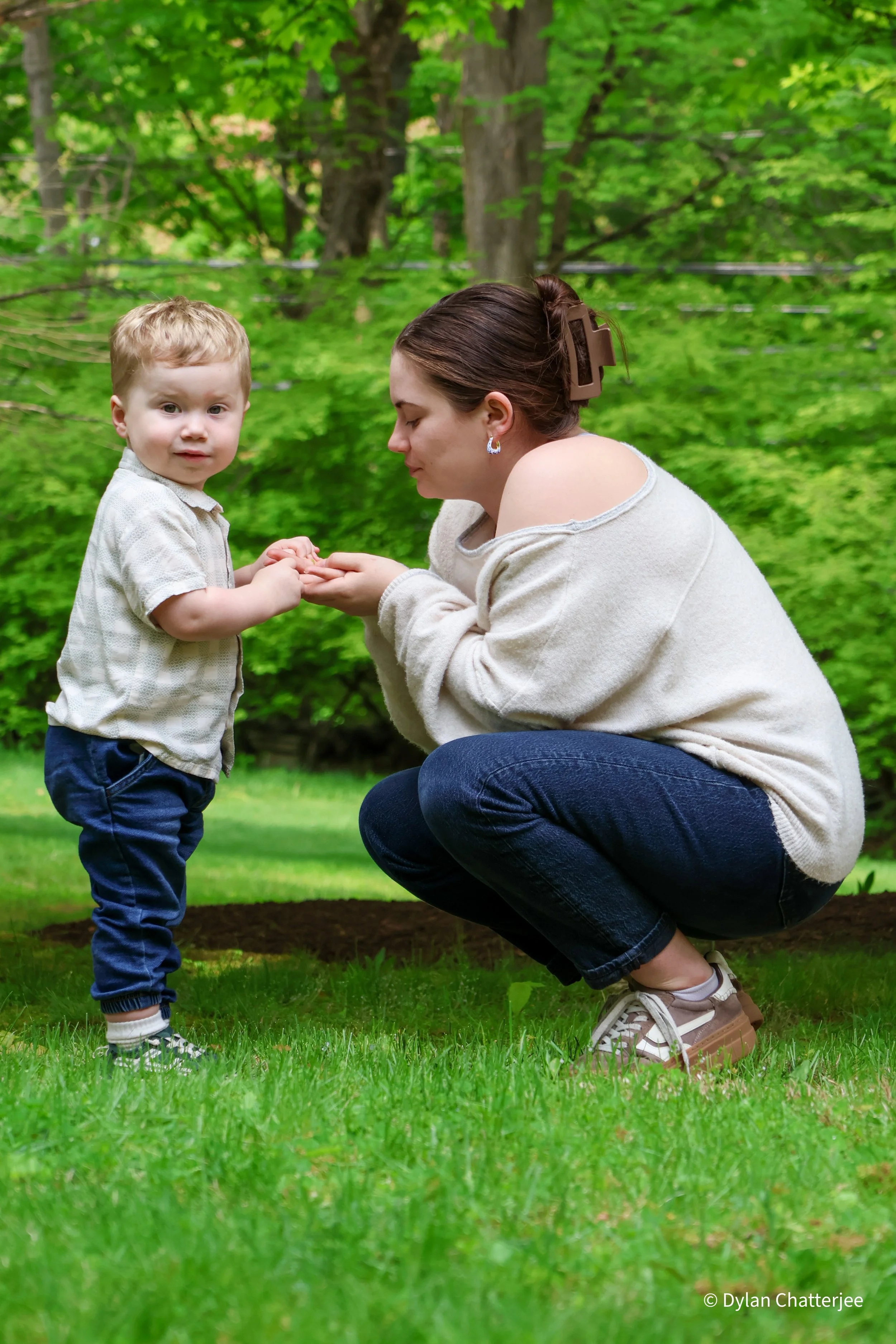 A woman kneeling on grass, holding hands with a young boy. The woman has her eyes closed and is wearing a beige sweater, jeans, and sneakers, with a large hair clip. The boy is standing, facing the camera, wearing a light-colored shirt, dark pants, a
