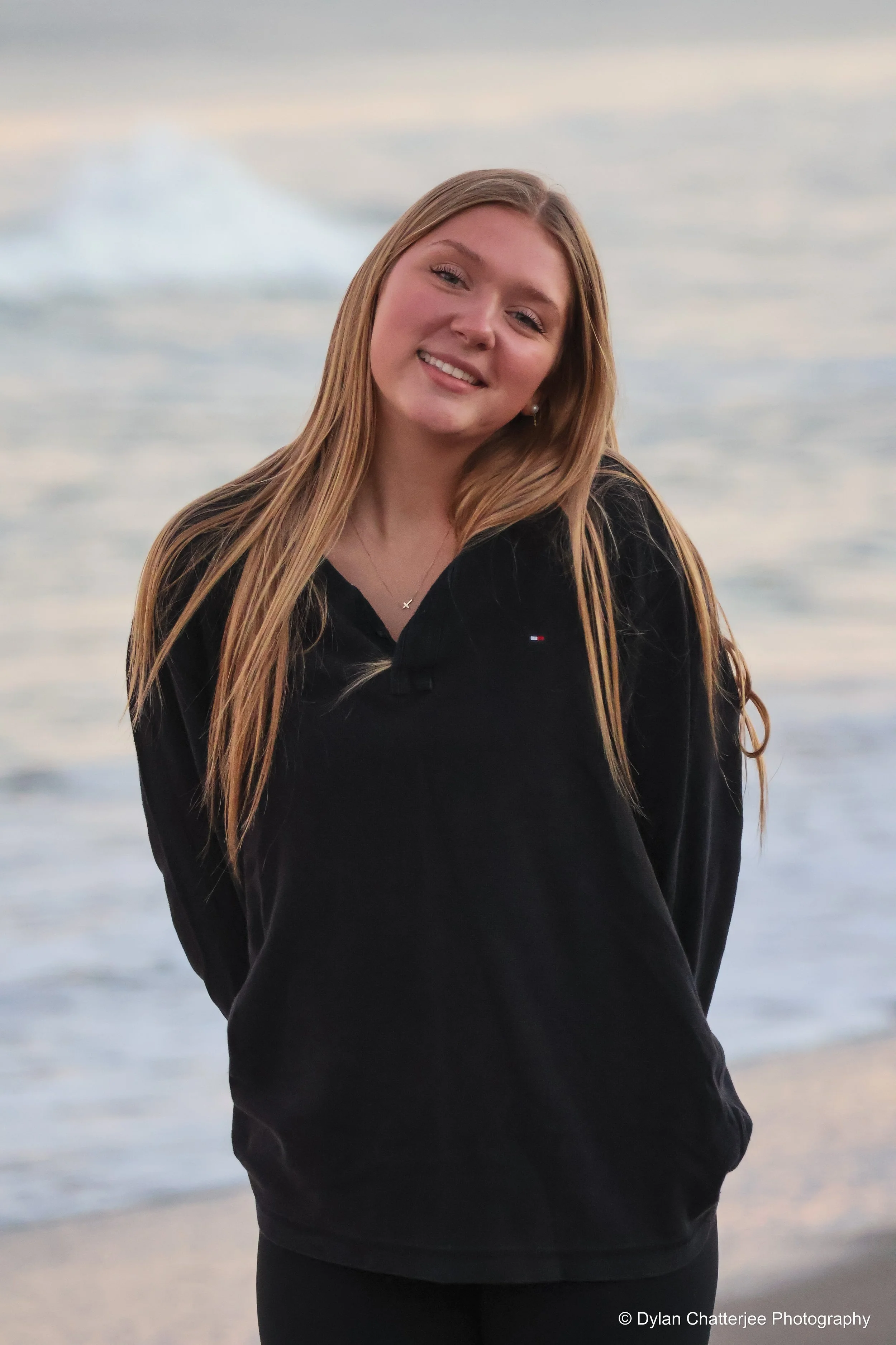 A young woman with long blonde hair smiling, standing outdoors before a blurred background of water and sky, wearing a black long-sleeve top and pearl earrings.