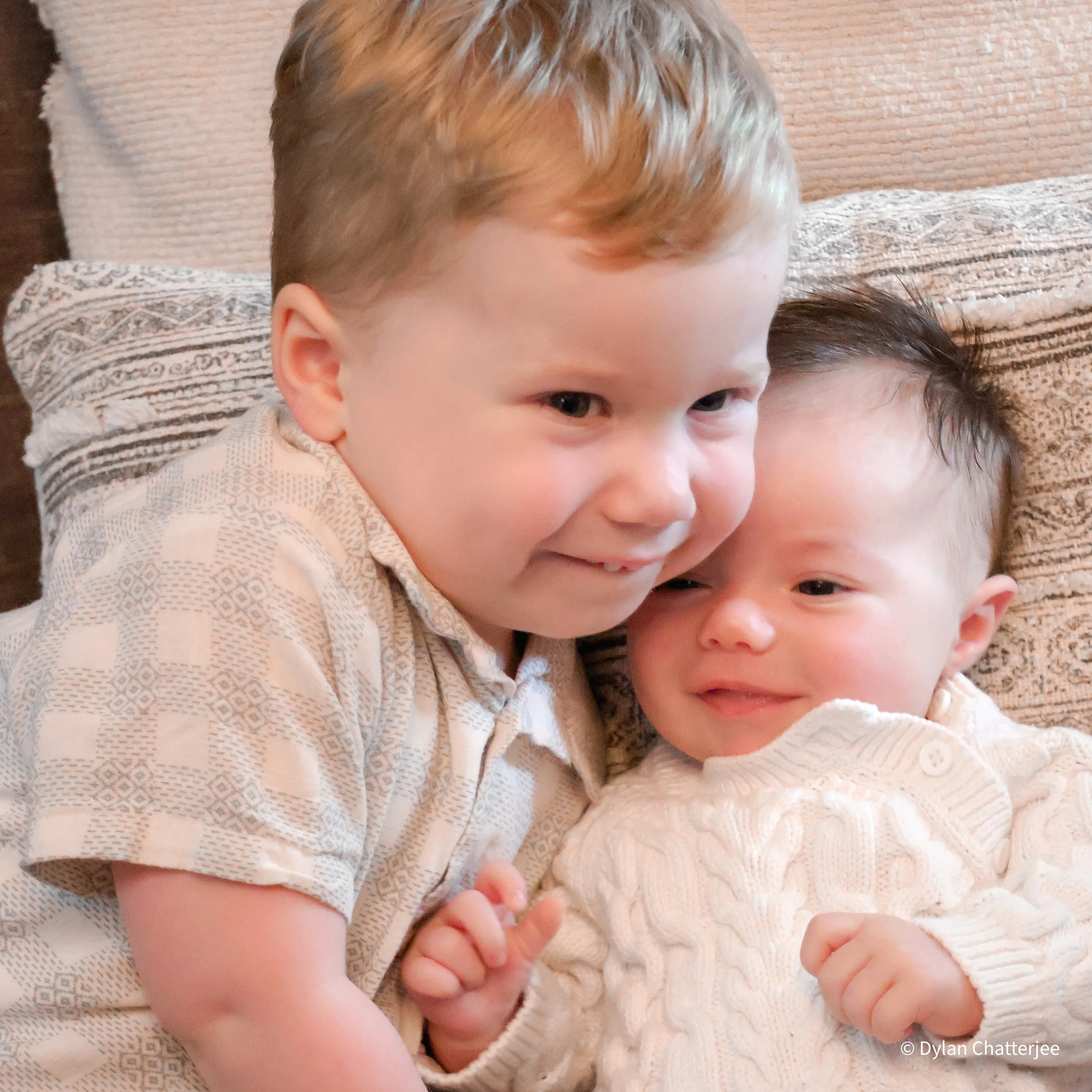 Two young children, a boy with light brown hair and a girl with dark brown hair, are lying close together on a beige and brown patterned blanket or couch, smiling and appearing happy.