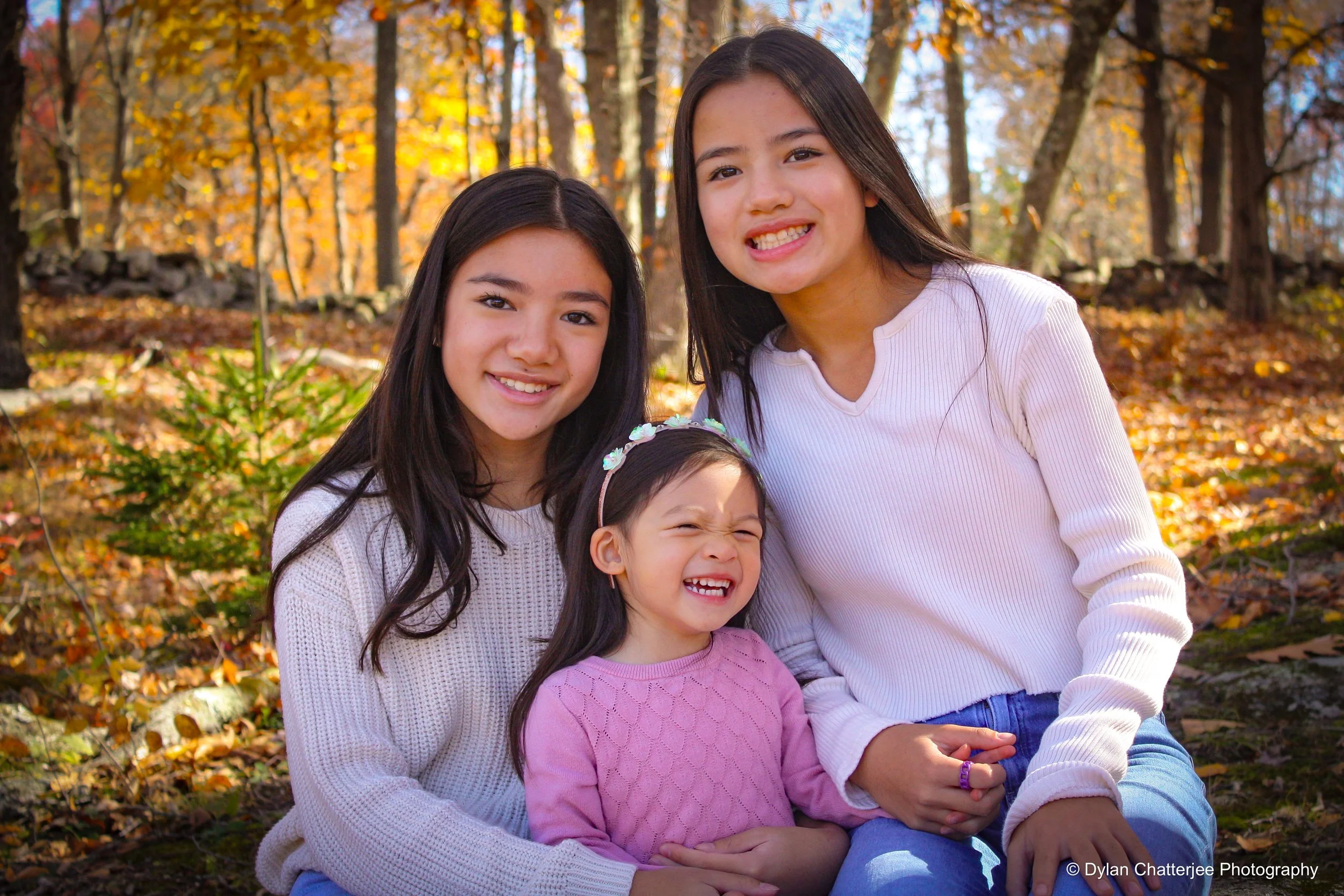 Four girls sitting outdoors in a park during autumn, smiling at the camera. The background has trees with yellow and orange leaves.