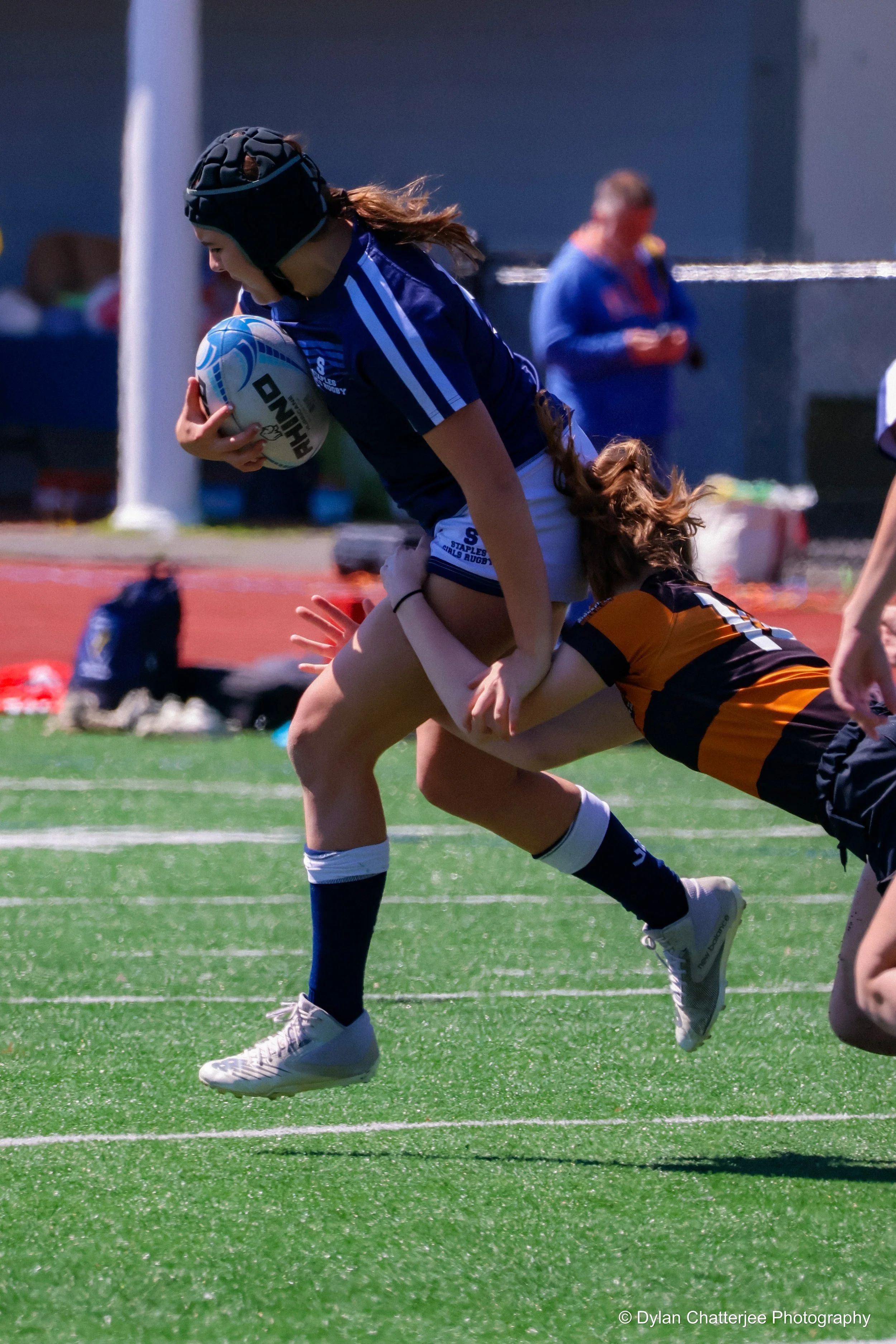 A female rugby player is running with a ball during a match, wearing a blue jersey and black protective headgear, while an opponent in a black and orange jersey attempts to tackle her on a green artificial turf field.