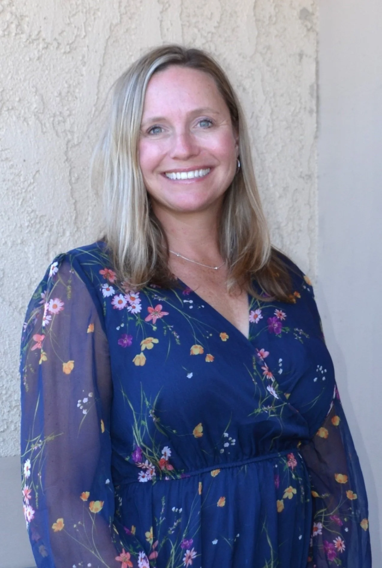 A woman with shoulder-length blonde hair smiling, wearing a blue floral dress, standing against a beige textured wall.
