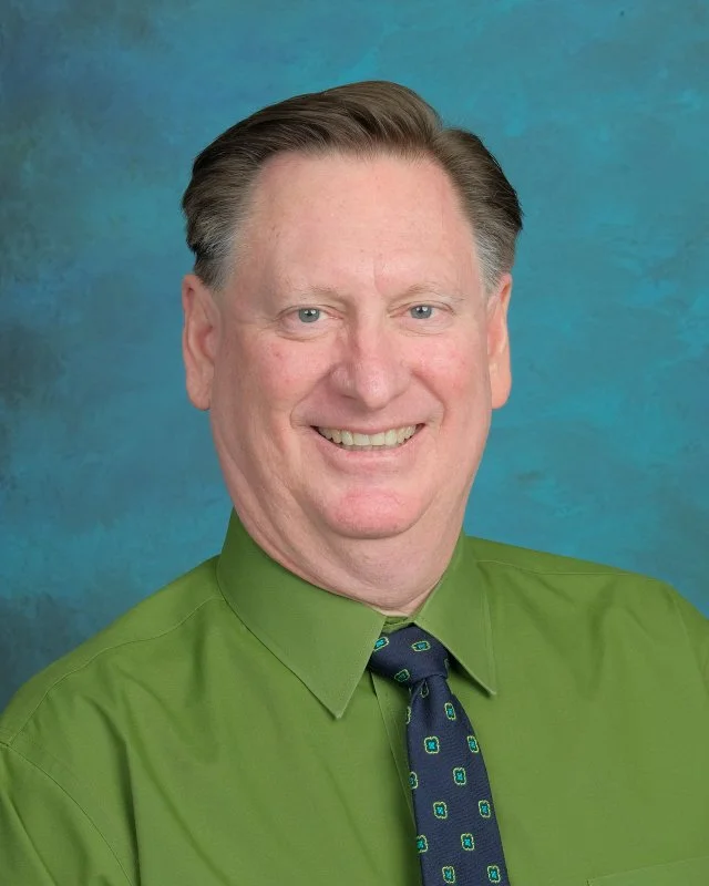 Headshot of a middle-aged man wearing a green shirt and a dark blue tie with small pattern, smiling against a blue textured background.