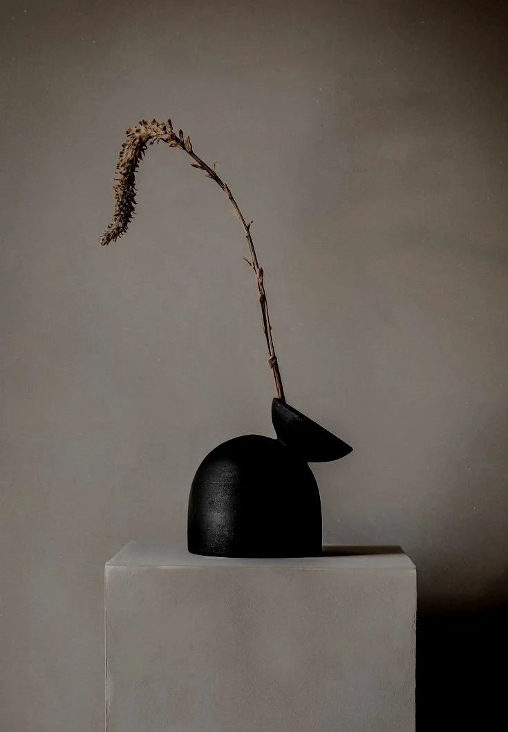 A minimalist black vase with an angled spout on a pedestal, holding a single dried twig against a neutral background.