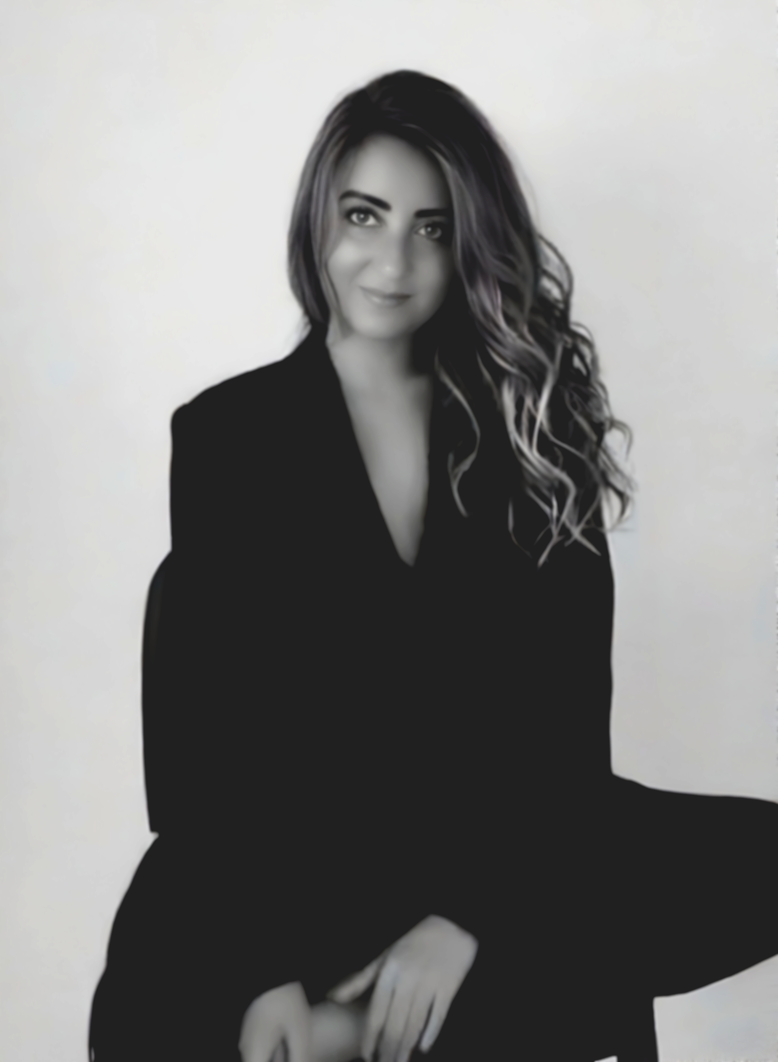 Black and white photo of a woman with long wavy hair, sitting in a relaxed pose, wearing a dark blazer and smiling subtly, against a plain light background.