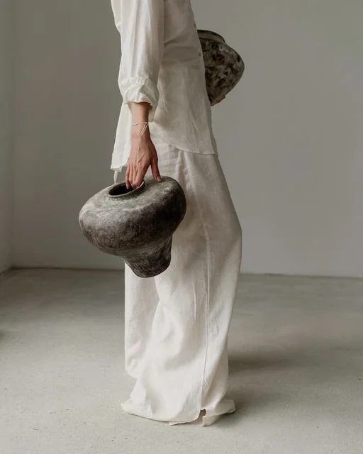 Person dressed in white holding two irregularly shaped stone vases, standing in a minimalistic room with plain white walls and concrete floor.