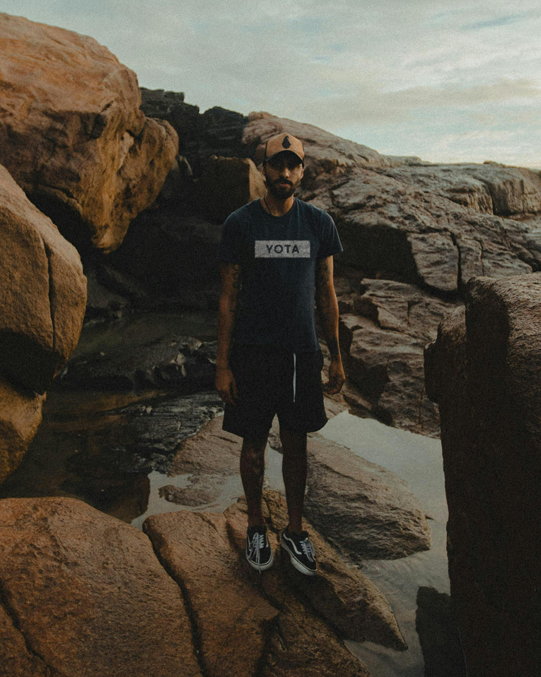 A man standing on rocks near water with a backdrop of a cloudy sky and rocky landscape