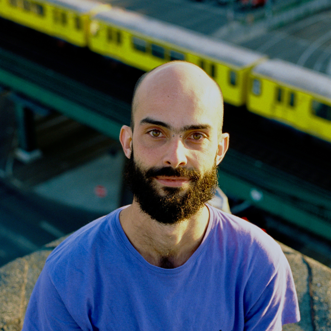 Close-up of a person with light skin and a dark beard, smiling, wearing a pink knit hat and a brown coat with a fuzzy collar, outdoors on sunny day with buildings and leafless trees in the background.