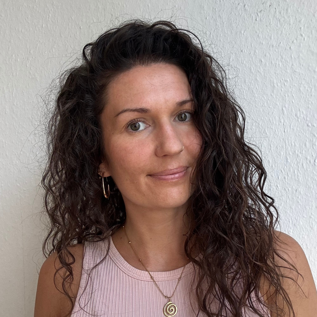 A woman with curly dark hair, wearing a pink sleeveless top, gold hoop earrings, a nose ring, and layered gold necklaces, standing against a plain white wall.