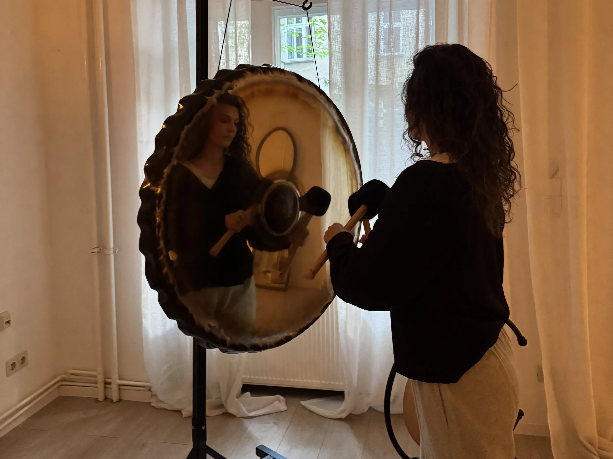 A woman with curly hair looking into a large, circular mirror with a black frame. She is in a room with a window and sheer curtains in the background. The woman appears to be holding a drumstick or mallet, with a partially visible object that looks like a percussion instrument in front of her.