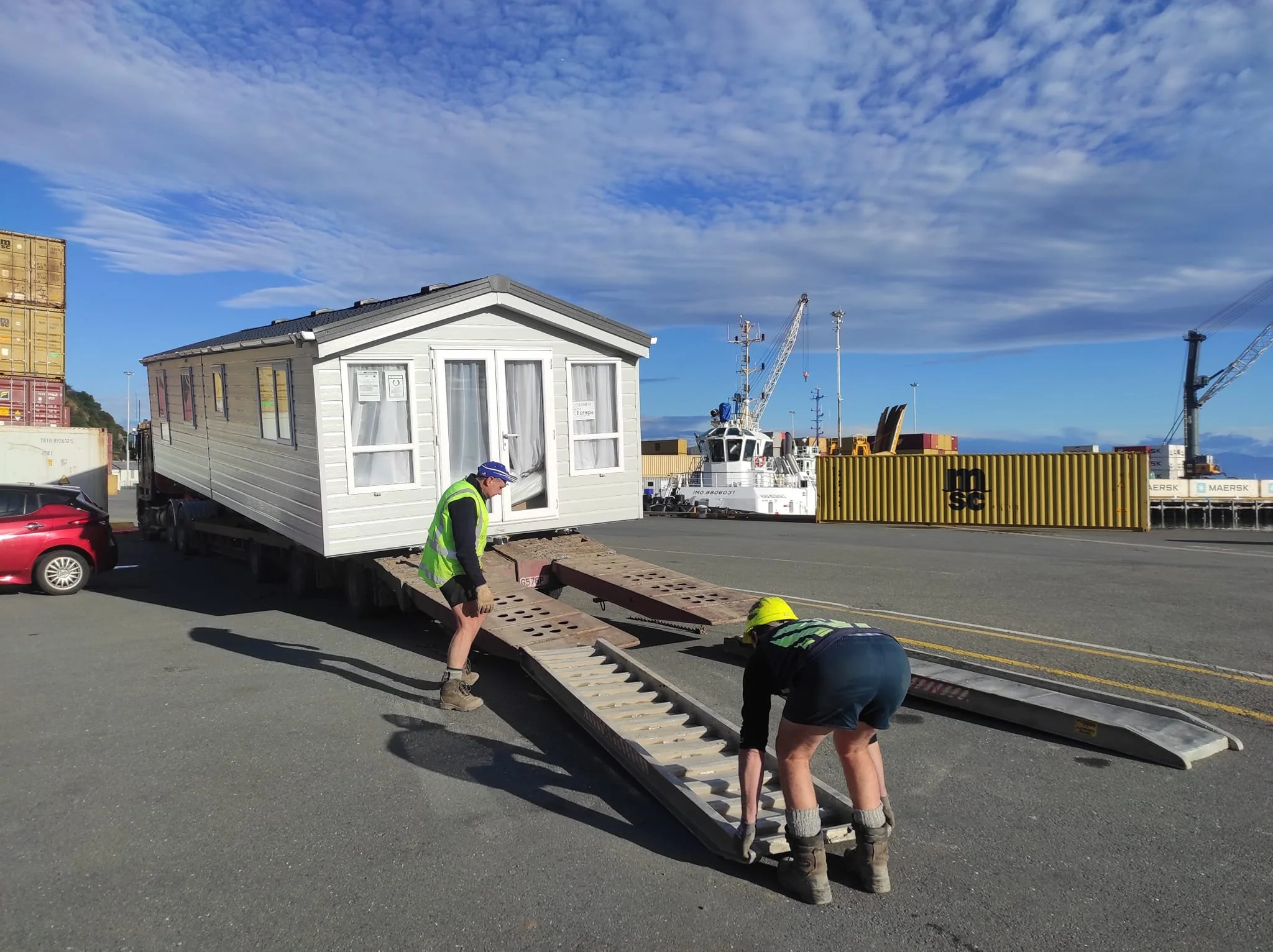 Two workers in safety vests and boots move a small house onto a truck trailer at a port near boats and shipping containers, under a partly cloudy sky.