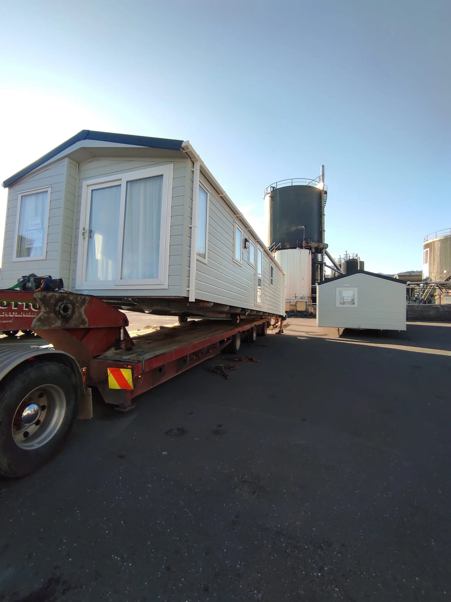Two mobile homes being transported on a flatbed trailer at an industrial site with large storage tanks in the background.
