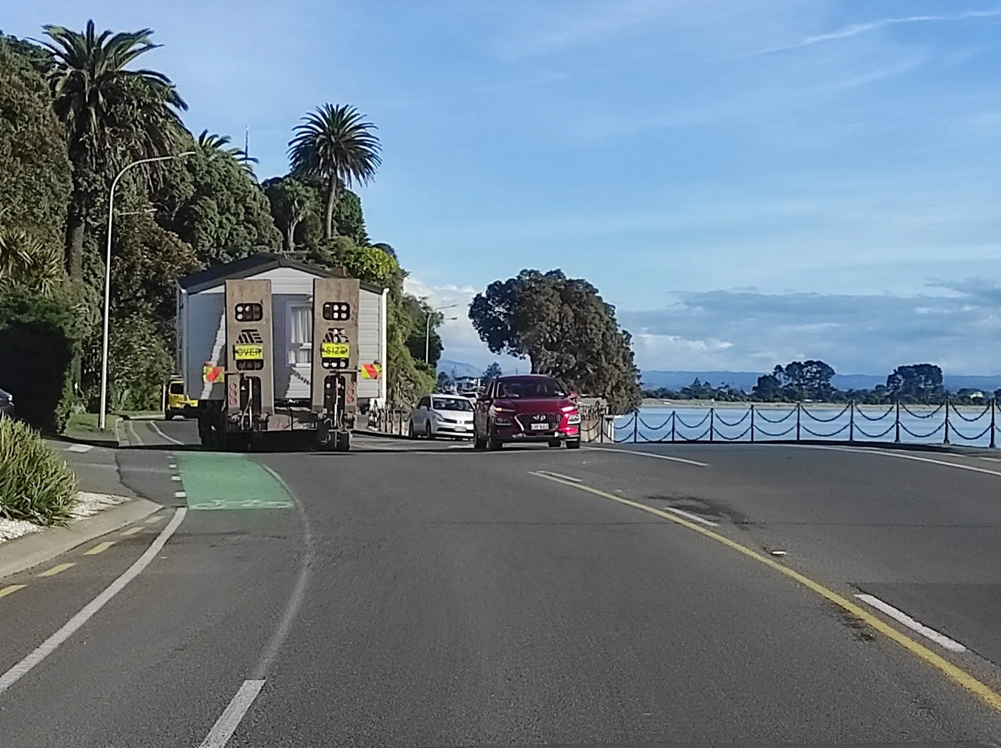 A scenic coastal road with palm trees on a hillside on the left, a small building on the back of a truck, and several cars driving along the road, with a large body of water and trees in the distance under a partly cloudy sky.