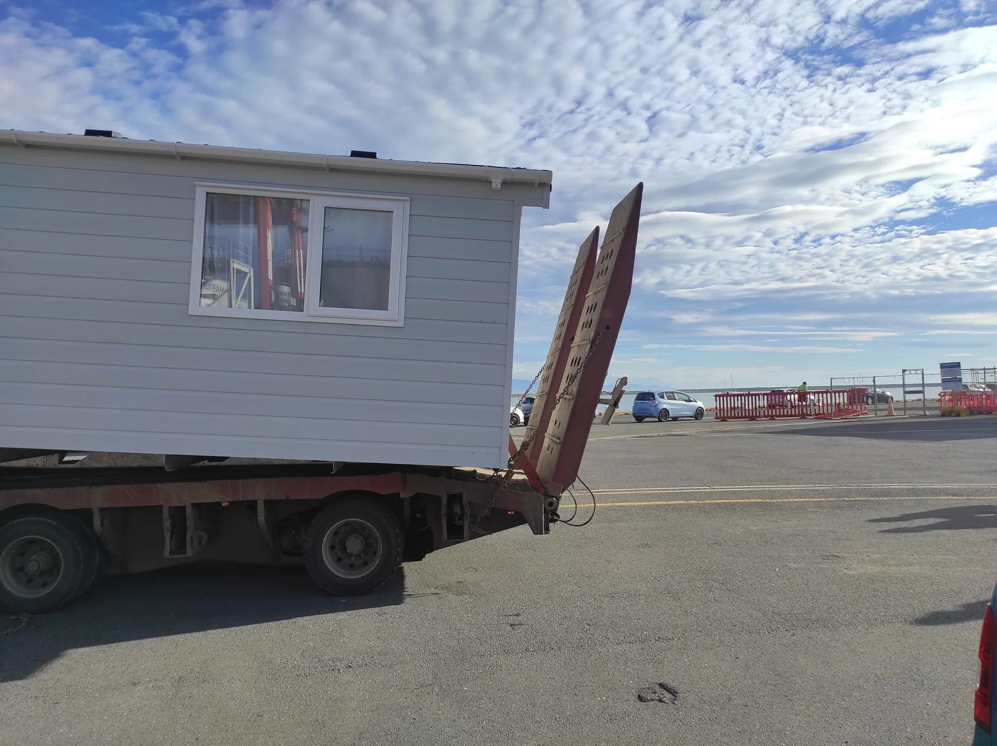 A white mobile home on a flatbed truck parked outdoors near a body of water with a cloudy sky and several cars in the background.