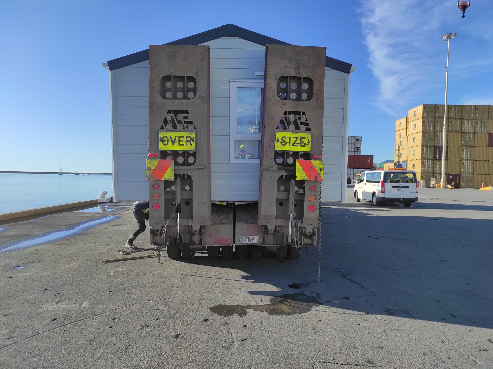 A truck with large concrete barriers on the back, marked with yellow signs 'OVER' and 'SIZE', parked on a concrete surface near water, with a white house in the background, a worker under the truck, and stacked shipping containers in the distance.