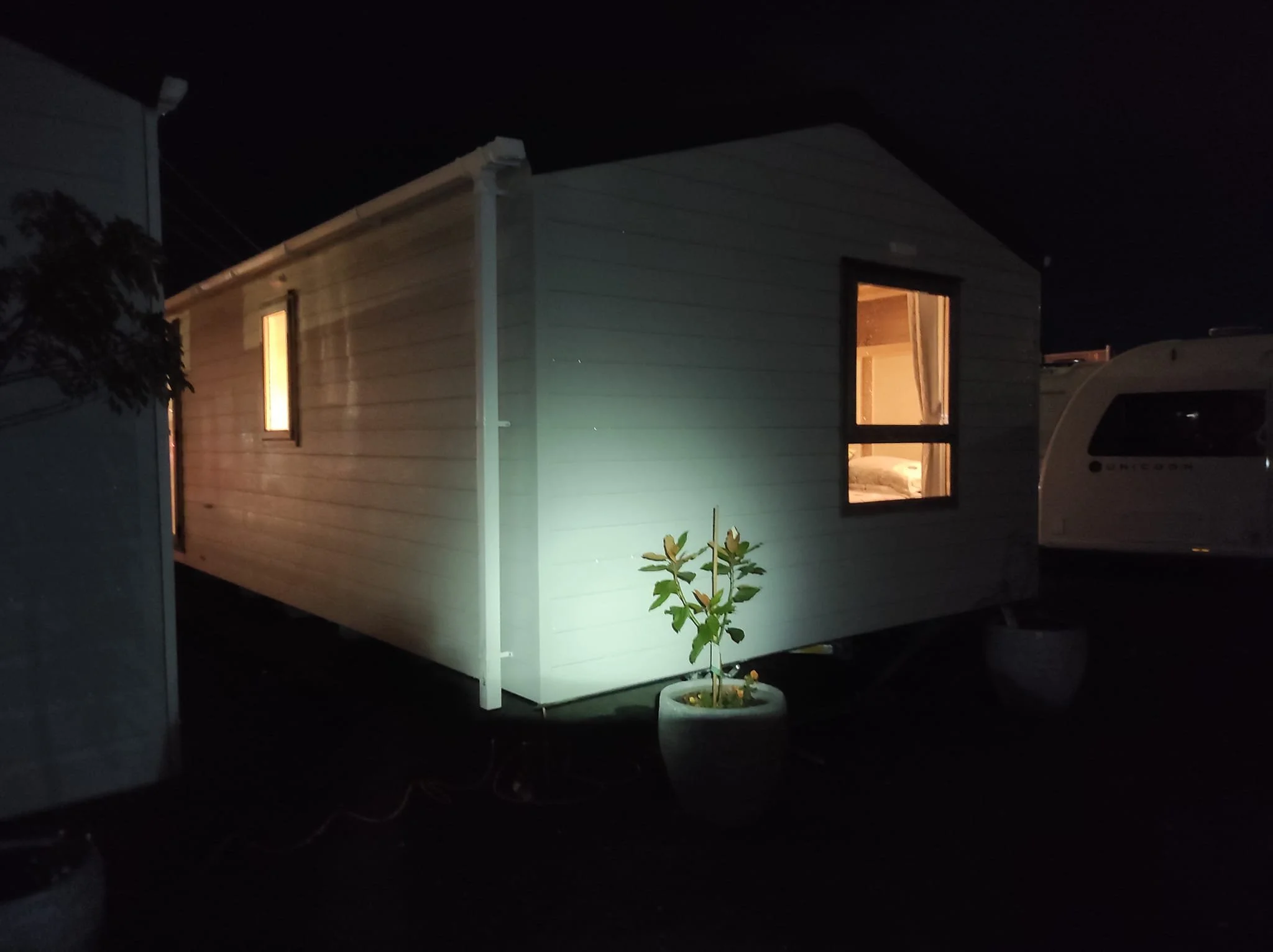 A white tiny house on wheels illuminated from within at night, with two windows showing warm interior lights, a potted plant in front, and a camper trailer parked nearby.