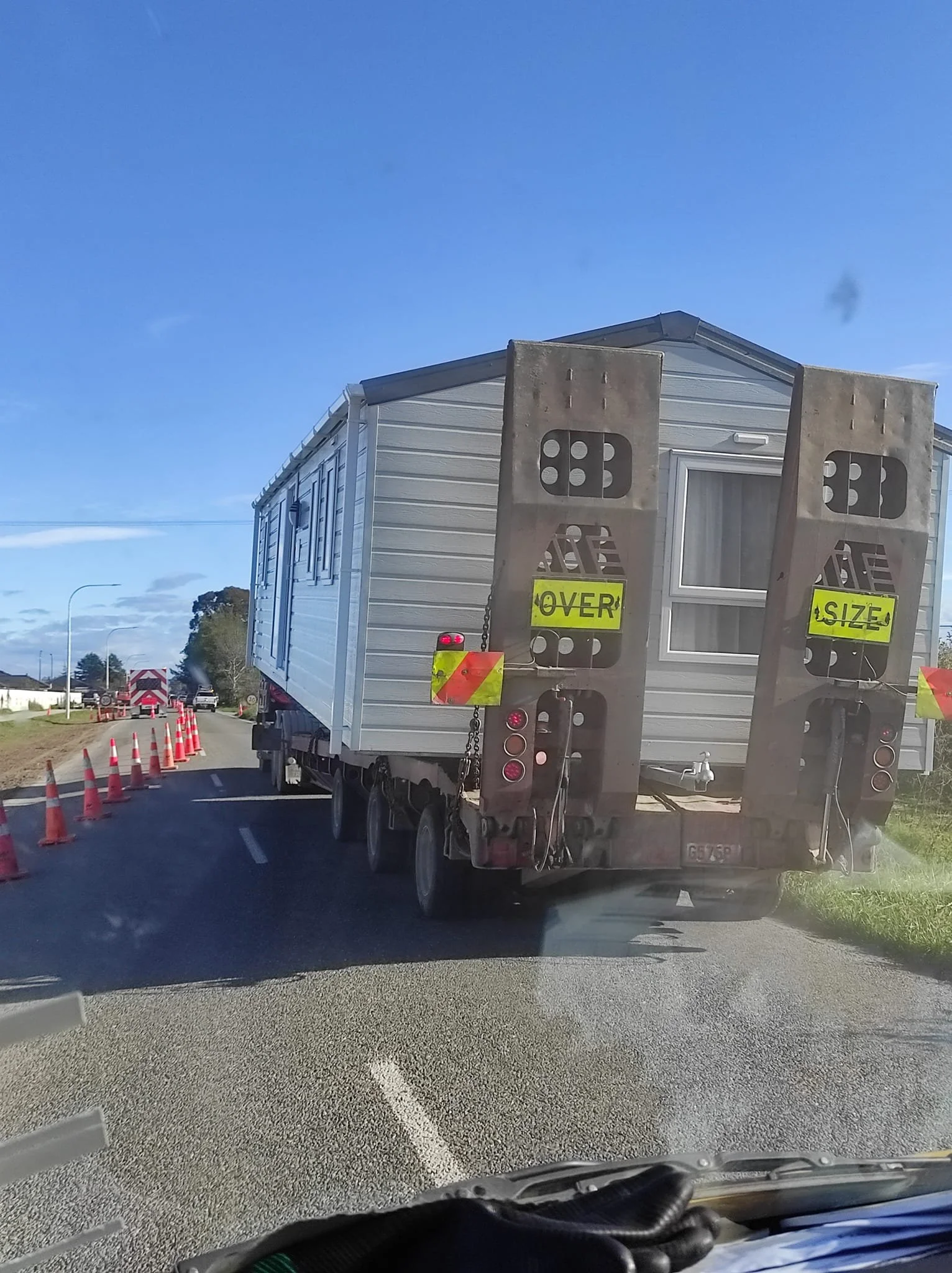 A truck carrying a tiny house on the road, with orange safety cones and a police vehicle in the background.