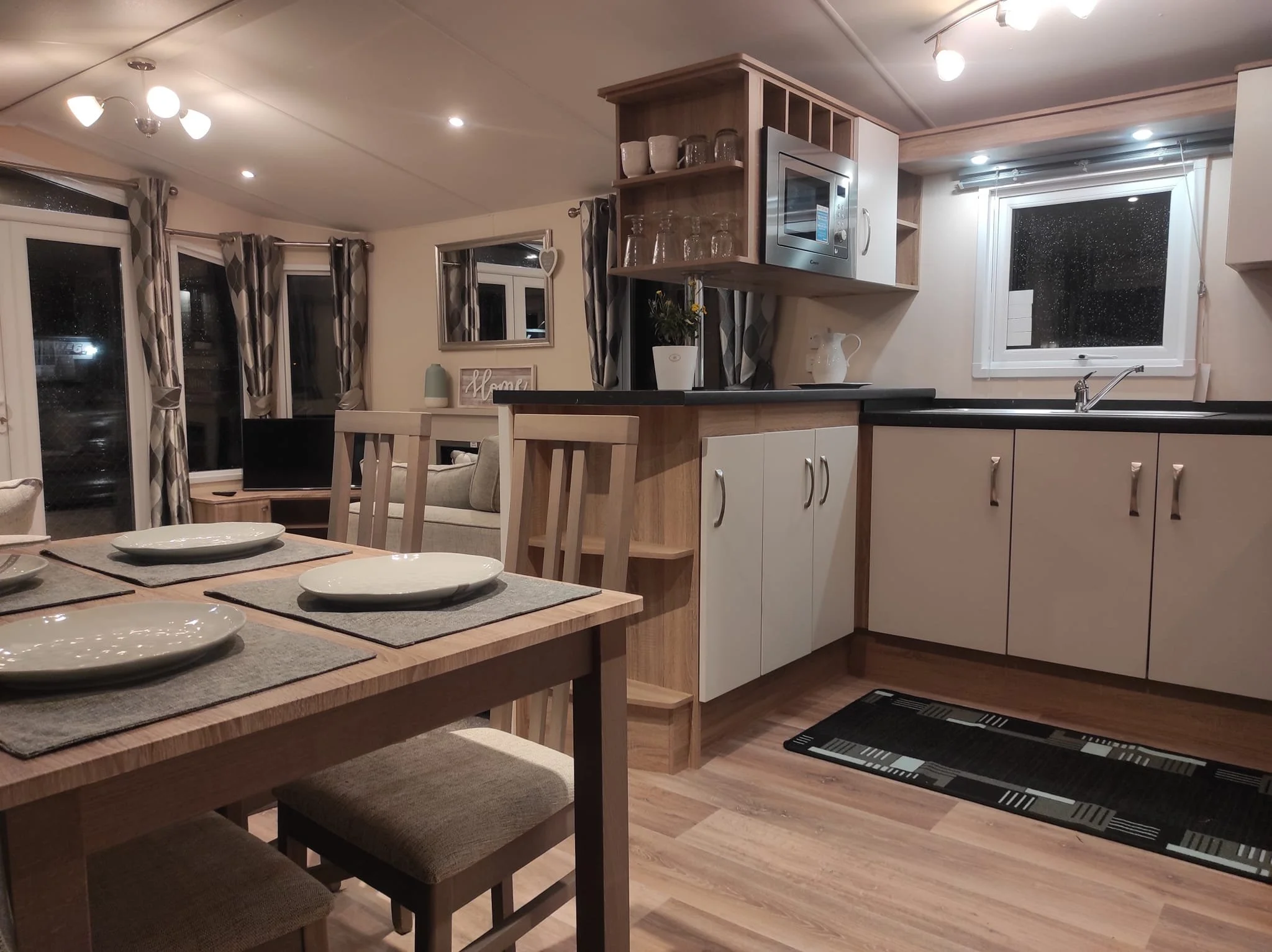 Interior view of a modern kitchen and dining area with a wooden dining table set for four, a living room with a television, and large windows with patterned curtains.