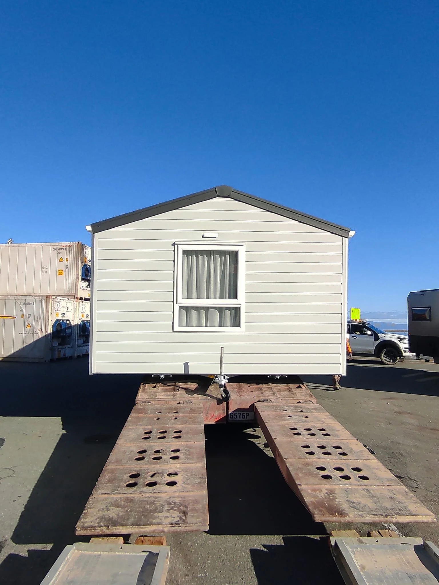 A small white house on a flatbed trailer with ramps, parked outdoors on a clear day with blue sky. There are vehicles and containers in the background.