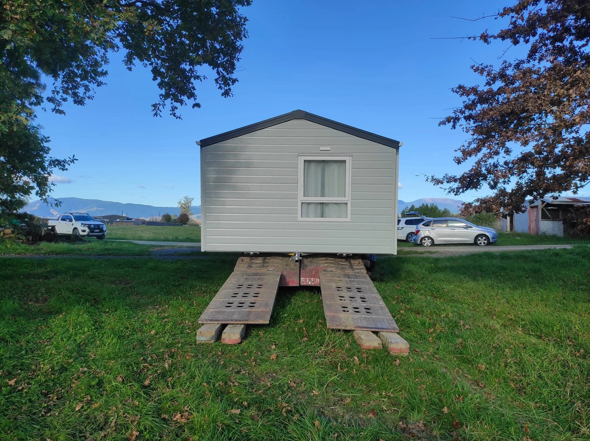 A small house on a trailer with wooden ramps in a grassy field, trees, distant mountains, and parked cars under a clear blue sky.