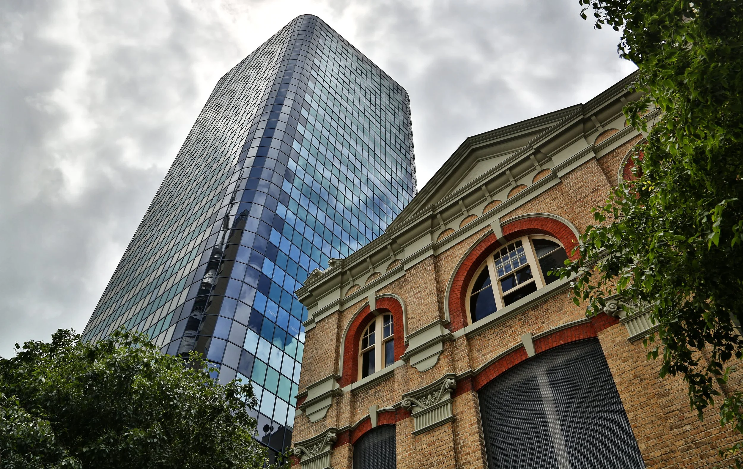 A low-angle view of a historic ornate brick building with arched windows standing in the foreground, with a towering modern glass skyscraper rising dramatically behind it against an overcast sky, framed by green trees on either side.