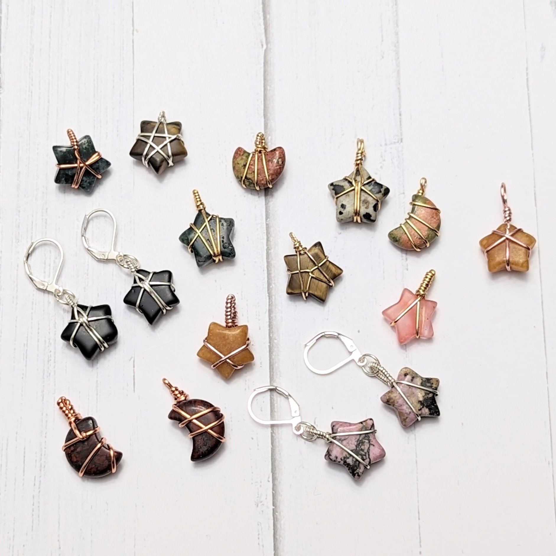 Various star-shaped and crescent-shaped gemstone jewelry pieces including earrings and pendants, with wire wrapping in gold, copper or silver, displayed on a white wooden surface.