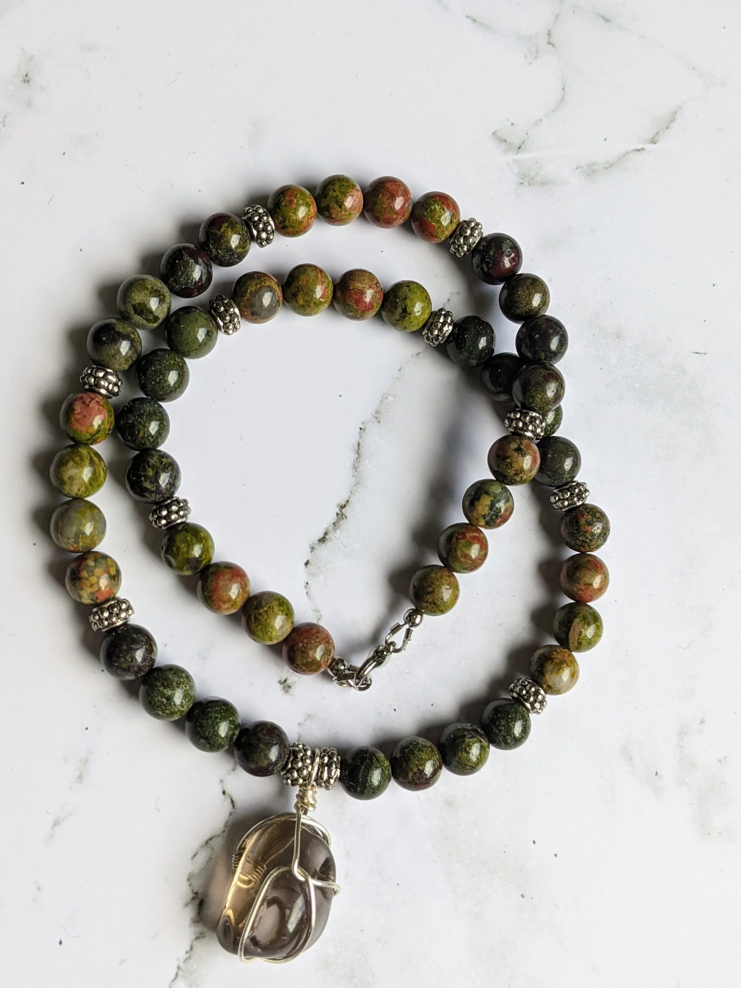 A beaded necklace with multicolored round stones and silver decorative spacers, featuring a large, oval-shaped stone pendant wrapped in wire.