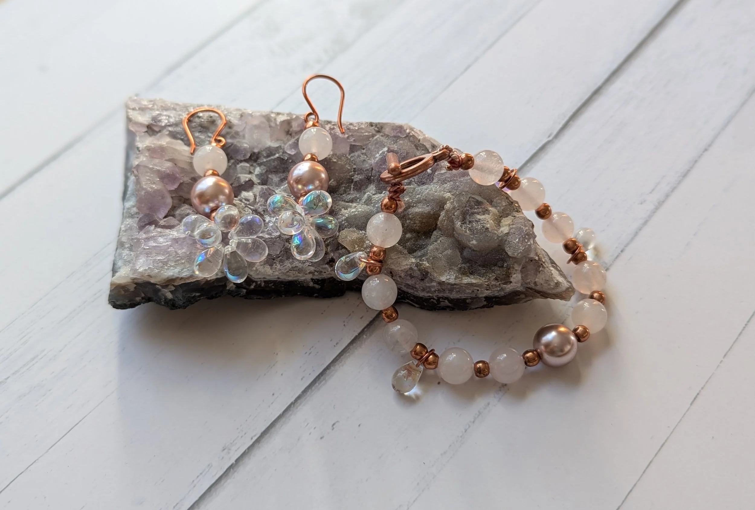Set of jewelry including pearl earrings, a pearl necklace, and a beaded bracelet with rose gold accents, placed on a slab of stone on a white wooden surface.