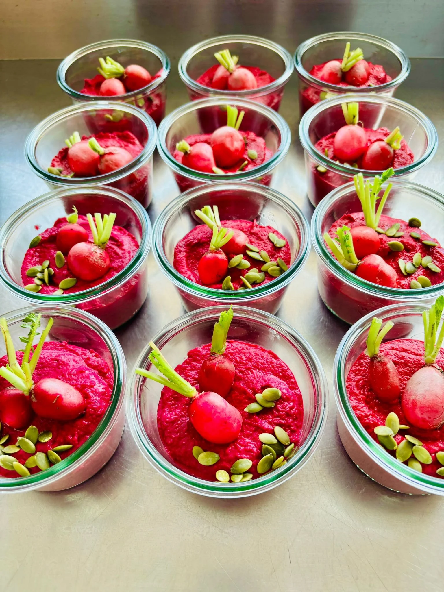 Glass cups filled with bright red beetroot mousse garnished with radishes and pumpkin seeds, placed on a metal surface.