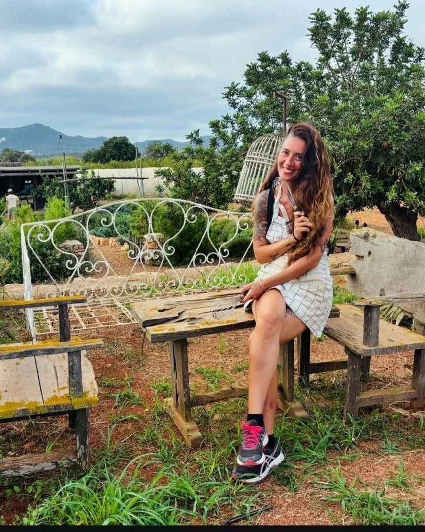 A smiling woman with long brown hair and tattoos on her arms, sitting on a weathered wooden bench outdoors in a garden or rural area, holding a pair of sunglasses.