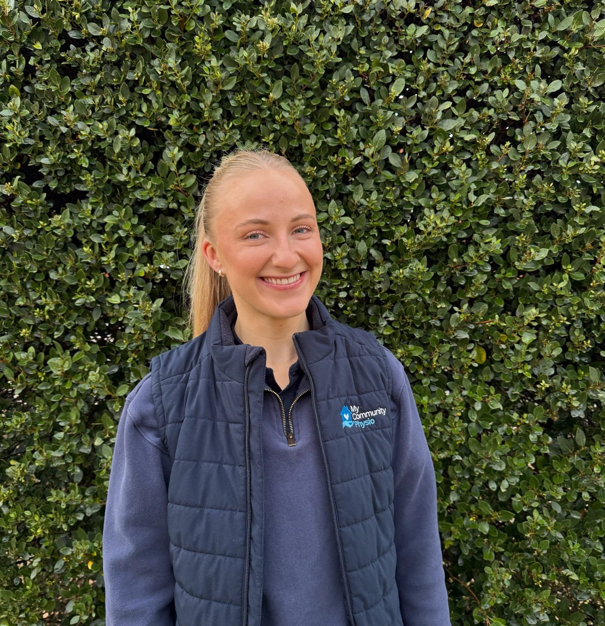 Close-up of a smiling woman with blonde hair wearing earrings and a dark blue jacket with a 'My Community Physio' logo, standing in front of a dense green shrub.