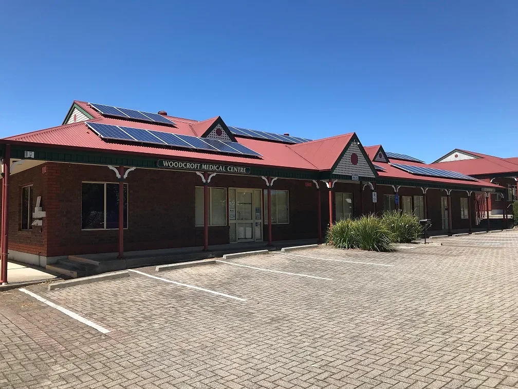 A brick building with a red roof and solar panels, labeled 'Woodcroft Medical Centre', with parking spaces in front and a clear blue sky.