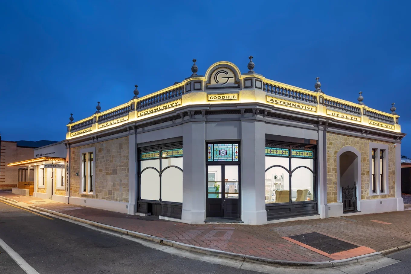 Corner building with illuminated signboards reading 'ALTERNATIVE HEALTH COMMUNITY' and 'GOODHUB,' featuring large stained glass windows on a street during dusk.