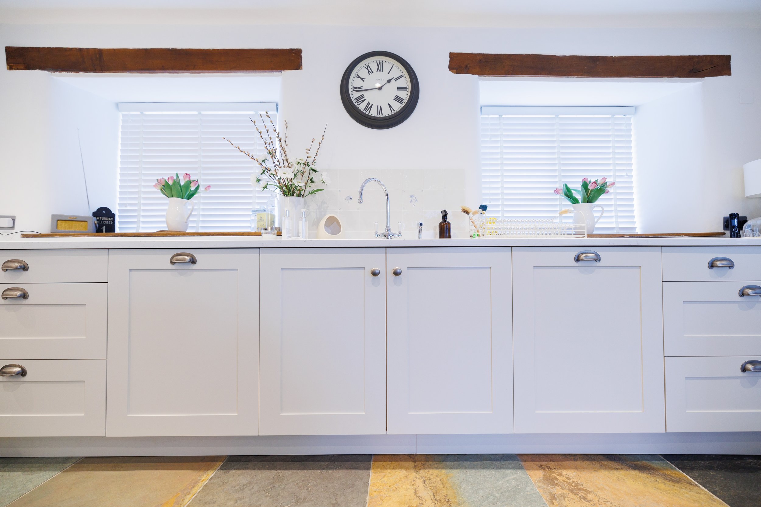 Kitchen with white cabinets, two windows with blinds, a black clock, and decorative vases with pink tulips.