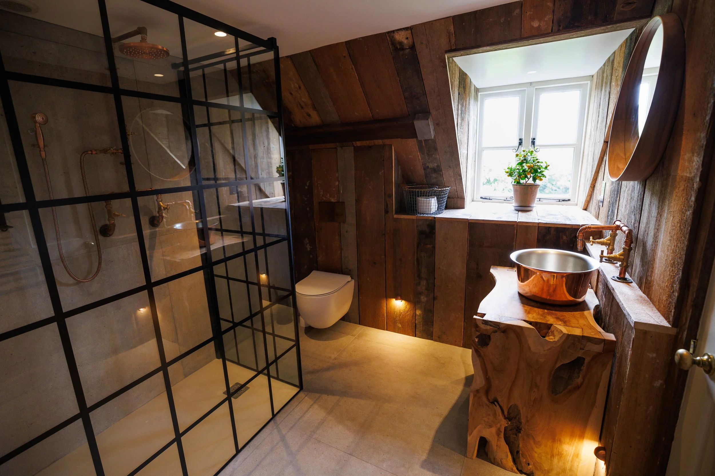A rustic bathroom with wooden walls and ceiling, a glass shower enclosure, a wall-mounted toilet, a small window with a potted plant, a wooden sink with a metallic bowl, and warm lighting.