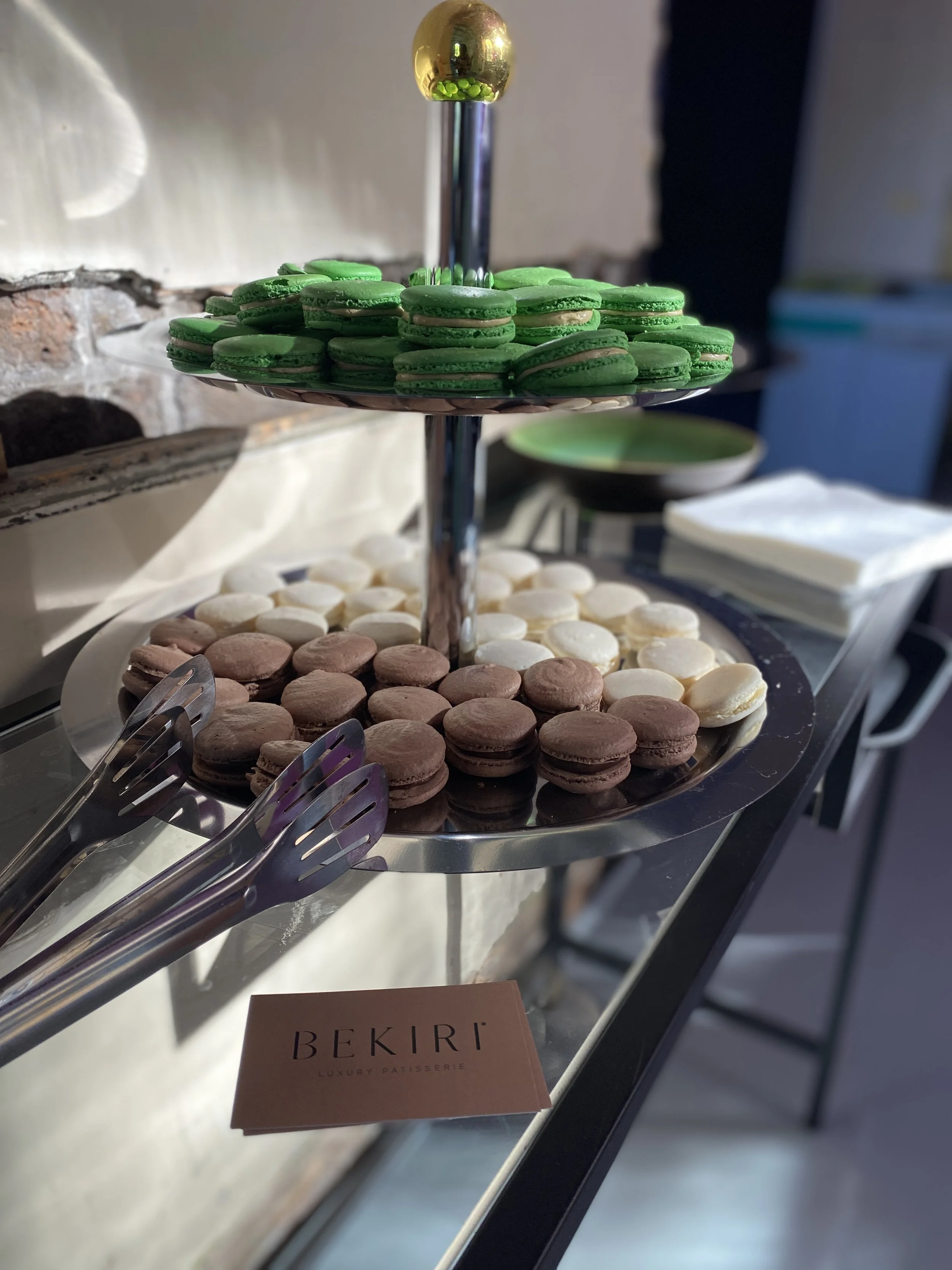 A two-tiered display stand holding an assortment of macarons in green, white, and brown. The macarons are arranged in a circular pattern on each tier. A small sign on the table reads 'BEKIRI LUXURY PATISSERIE'.