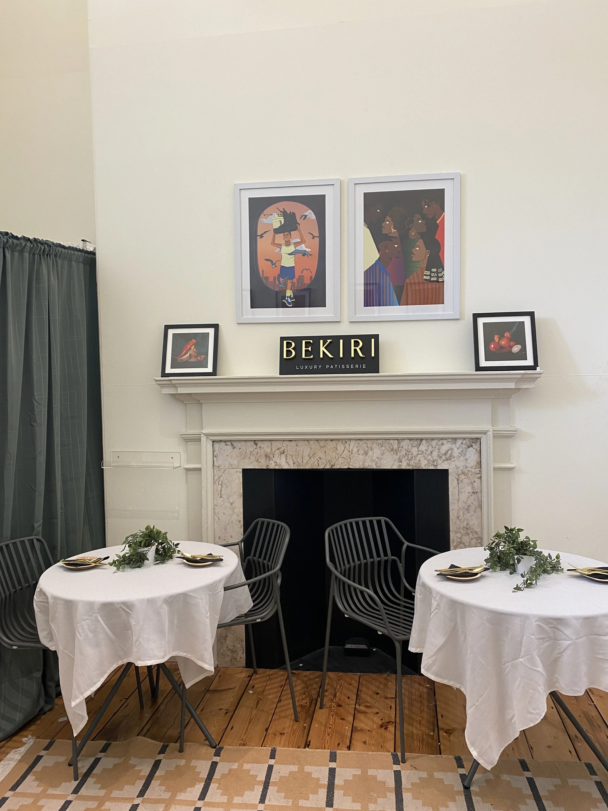 Interior of a dining area with two round tables covered with white tablecloths, decorated with green foliage, set with gold and black plates and cutlery. There are four black chairs around the tables. Behind, a fireplace with a marble surround and a beige wall above displays framed artwork: two illustrations above and two smaller framed photos of tomatoes and a bird. A sign in the center reads 'BEKIRI LUXURY PATISSERIE'.