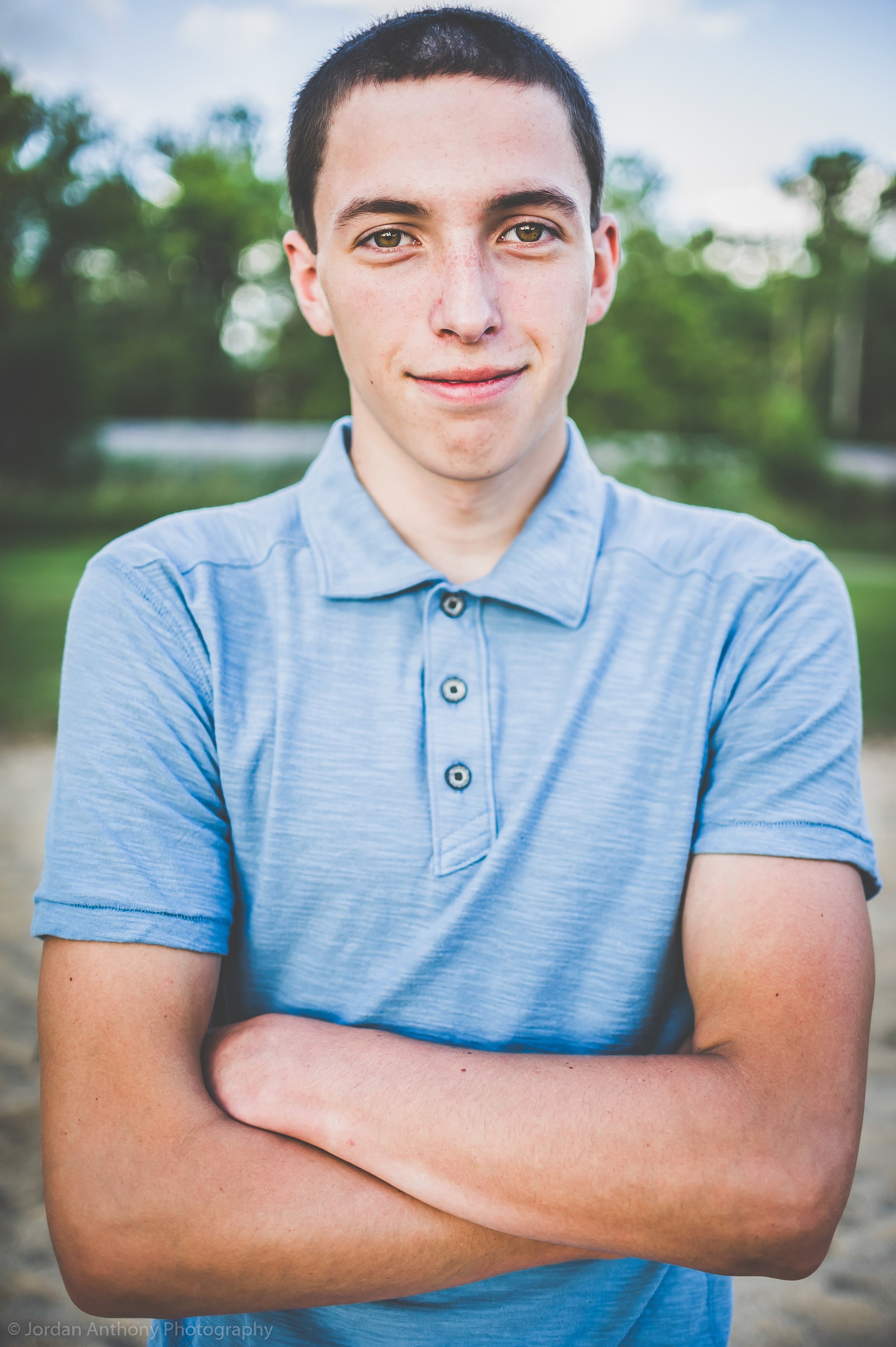 A young man with short dark hair, wearing a light blue polo shirt, standing outdoors with crossed arms, smiling slightly with trees and sky in the background.