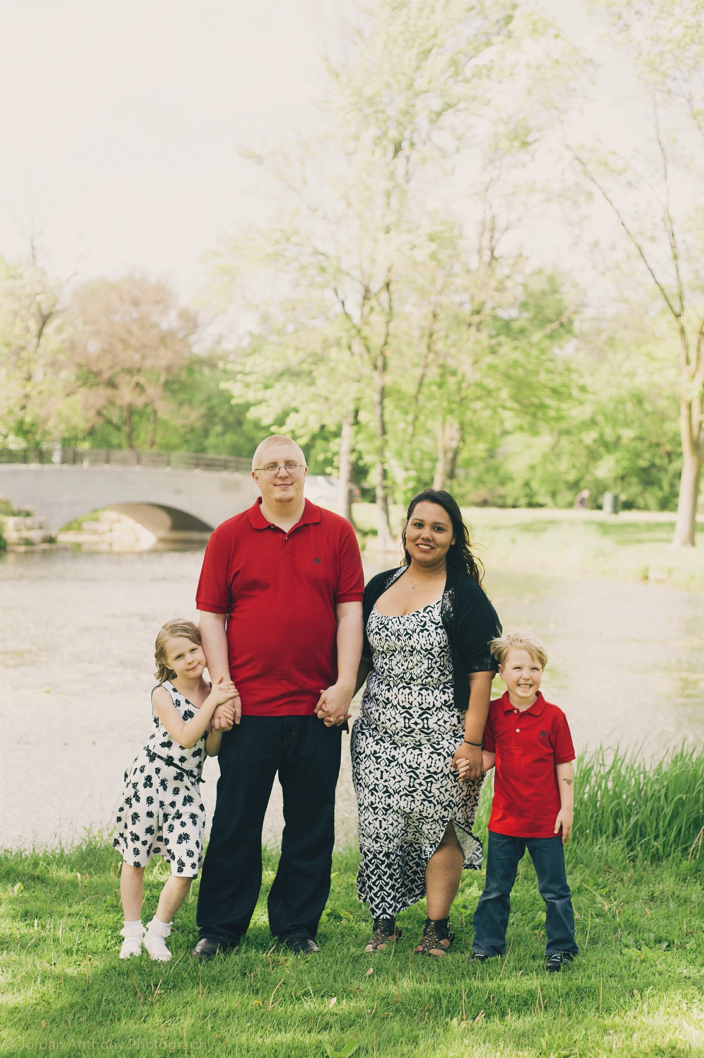 A family of four standing on grass near a pond with trees and a bridge in the background during daytime.