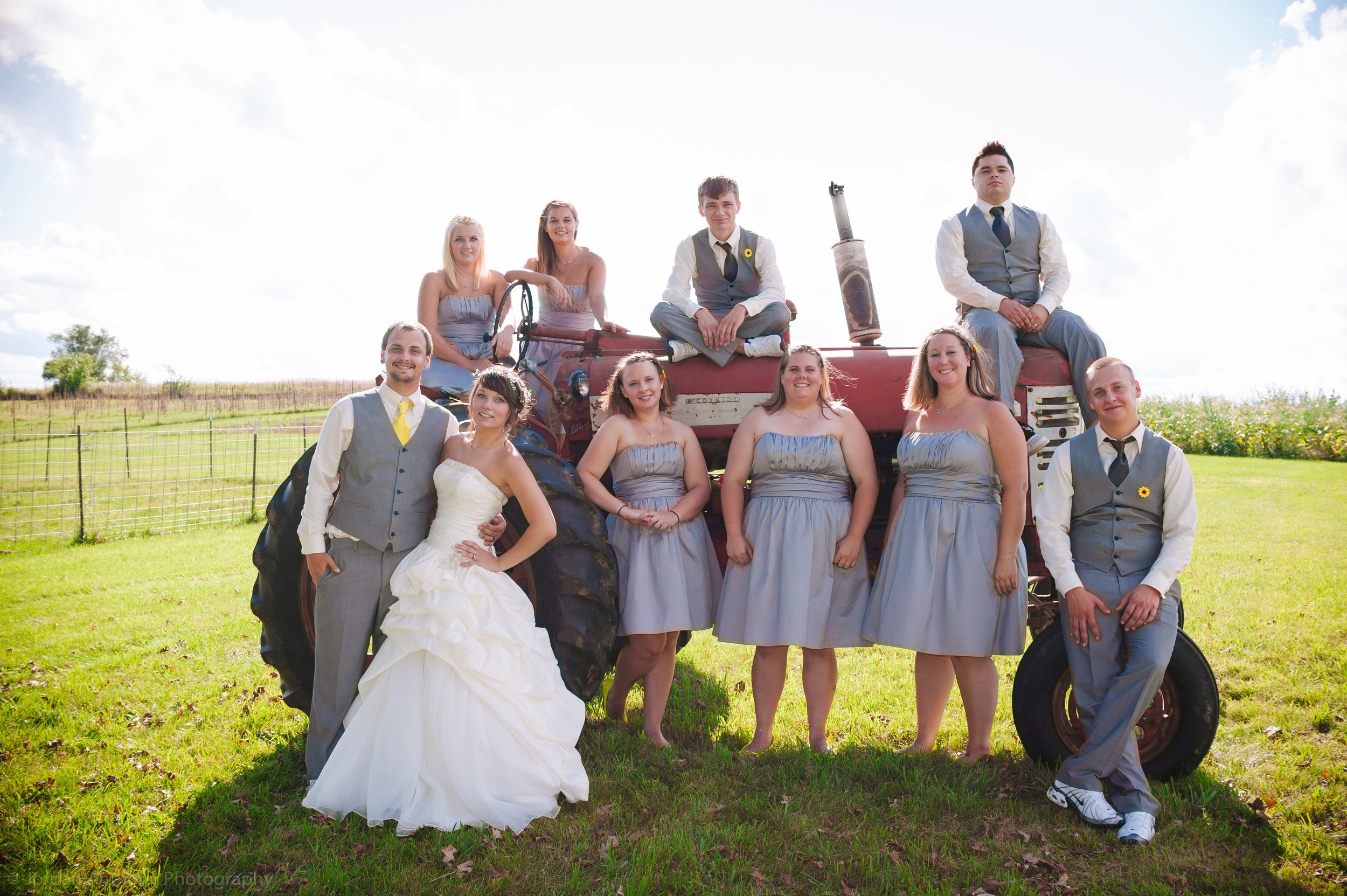 Group of people, including a bride in a white wedding gown and groom in a gray suit, posing on and around a vintage tractor in an open grassy field on a sunny day.