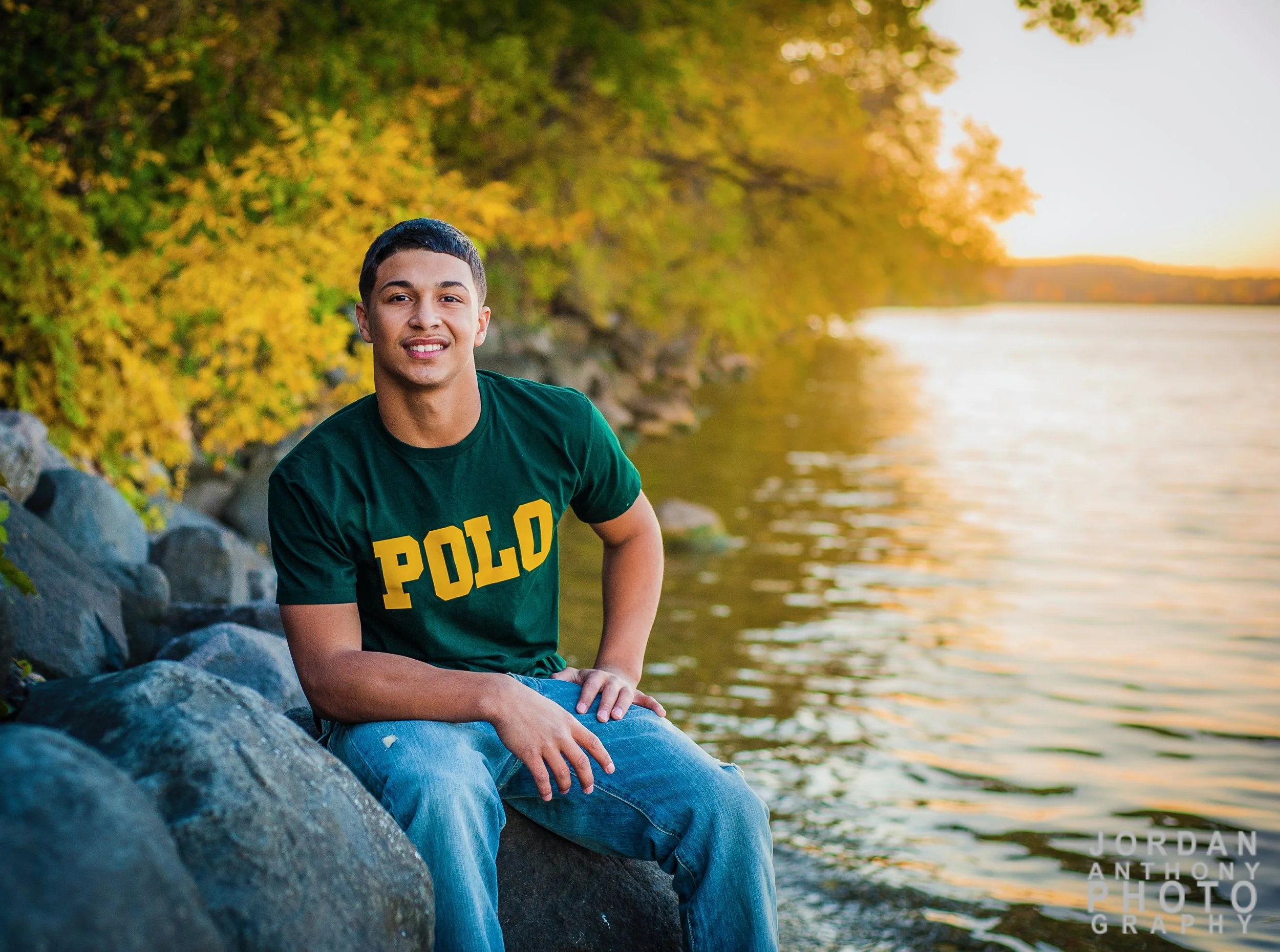 A young man sitting on rocks by a body of water with trees with yellow and green leaves in the background during sunset, wearing a green Polo shirt and jeans.