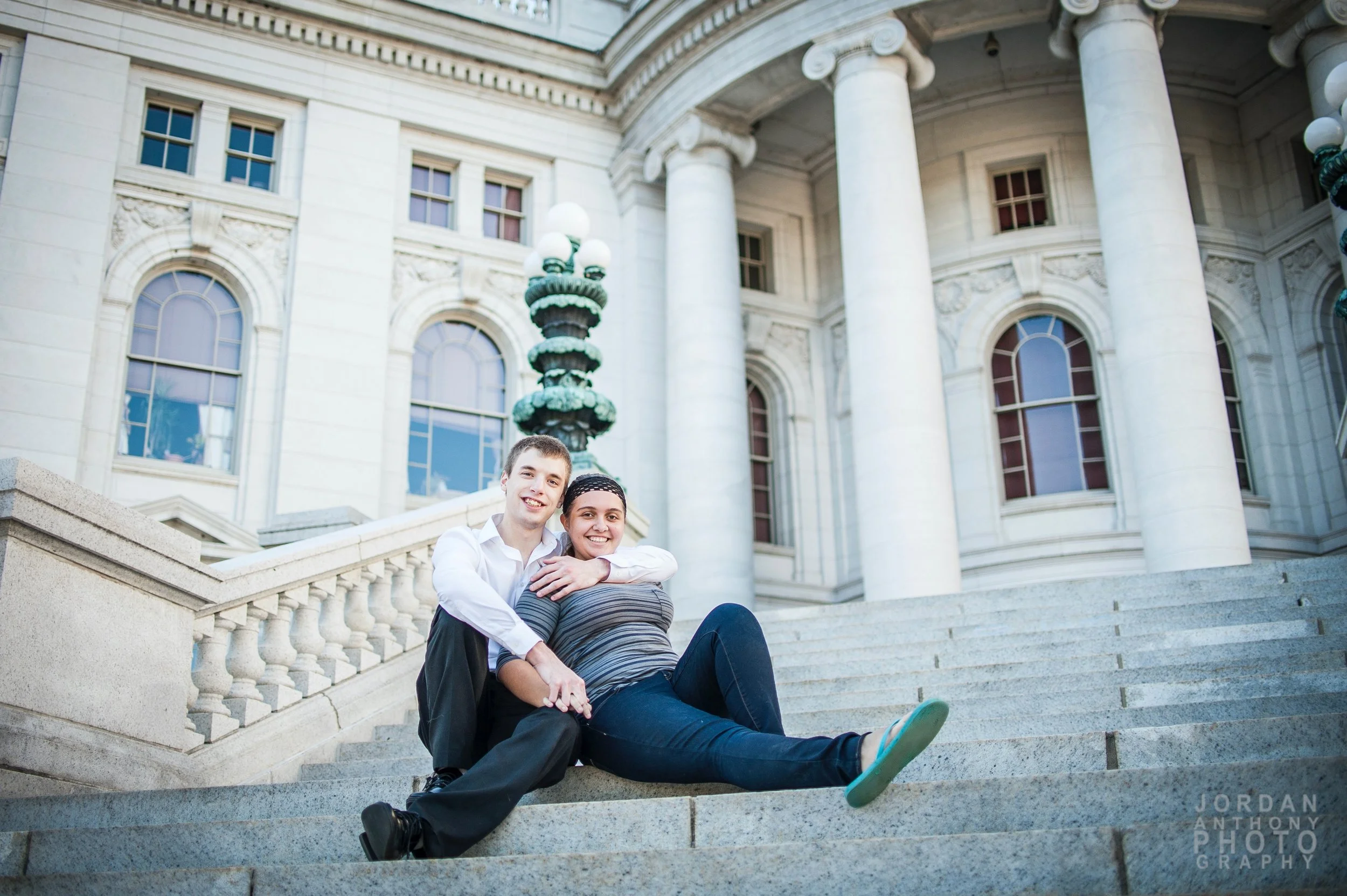 A young man and woman sitting on steps in front of a historic white building with large columns and arched windows, smiling at the camera.