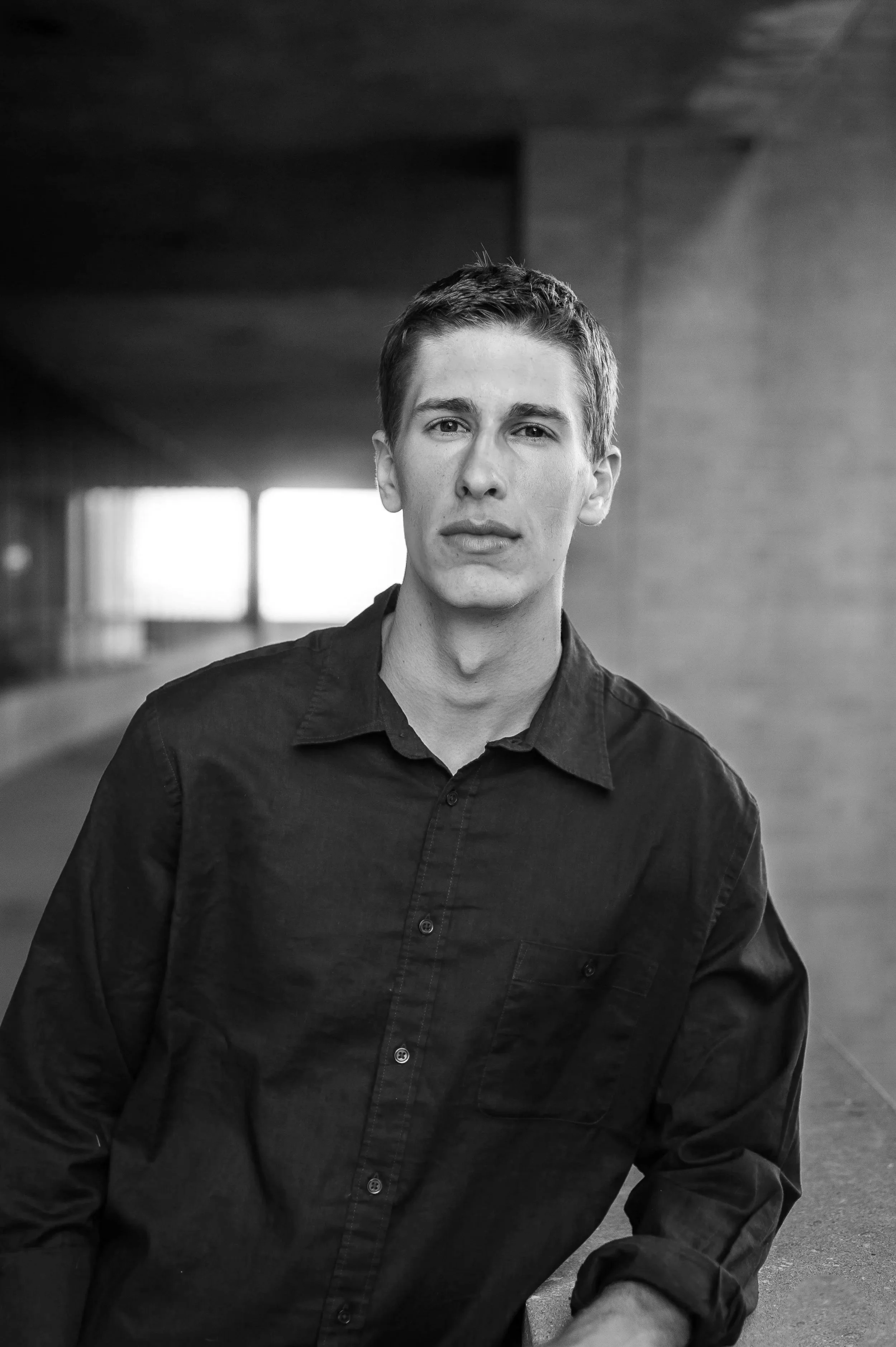 Black and white portrait of a young man with short hair, wearing a dark button-up shirt, standing in an urban environment with windows and concrete walls in the background.